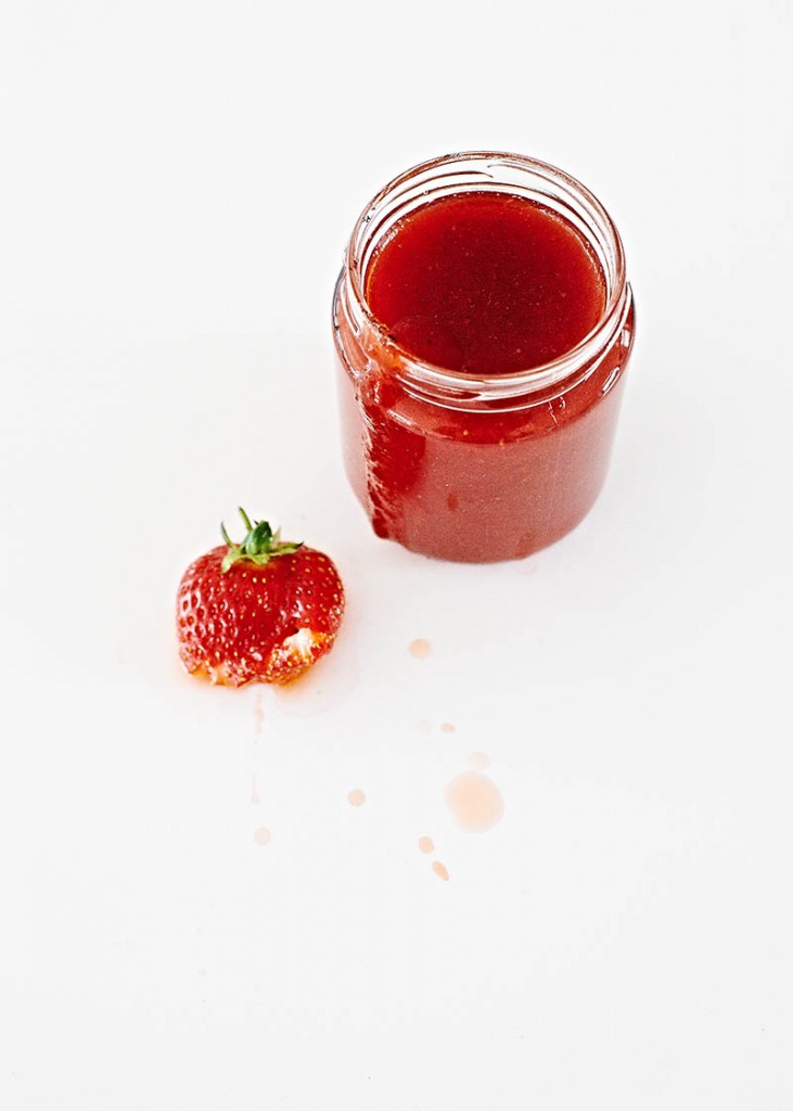 Ein Glas frisch gekochte Erdbeer-Marmelade und eine zerdrückte Erdbeere - plakatives und modernes Food-Motiv auf weißem Untergrund mit viel Freiraum